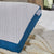 A close-up of the La-Z-Boy Down Alternative Pillow with a white and blue moisture-wicking, textured cover and branded tag, resting on a bed with a cream knitted blanket and white sheets. Sunlight streams in through the window.