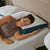 A woman with brown hair in a sleeveless taupe top lies on a bed with white textured bedding, resting her head on the La-Z-Boy Down Alternative Pillow by La-Z-Boy, featuring a white and teal cover—ideal for side sleepers.
