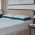 A modern bedroom features a beige upholstered headboard, two La-Z-Boy Down Alternative Pillows by La-Z-Boy with teal sides, a white textured mattress, beige bed skirt, and a round black nightstand with books.