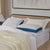 A neatly made bed features two La-Z-Boy Down Alternative Pillows with textured blue sides and white tops, stacked on white sheets with a cream knitted blanket. Beige curtains and a modern painting add to the stylish scene.