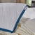 A close-up of the La-Z-Boy Serene Foam Hybrid Pillow shows its white textured surface with blue trim, resting on a cream blanket beside white sheets. A small tag displays the La-Z-Boy brand. Greenery is visible through a window in the background.