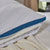 A close-up of the La-Z-Boy Serene Foam Hybrid Pillow highlights its white, textured, moisture-wicking cover with small blue patterns and a blue mesh edge. Partially unzipped, it reveals its inner layers as it rests on a cream knit blanket and light bedsheet.