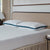 A neatly made bed with a beige upholstered headboard showcases two La-Z-Boy Serene Foam Hybrid Pillows with blue-green edges and a white textured mattress. A nightstand with books and a lamp is partially visible, framed by beige curtains at the window.