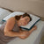 A woman with brown hair sleeps on her side, resting on a La-Z-Boy Serene Foam Hybrid Pillow with a moisture-wicking cover. She wears a sleeveless beige top and lies on a white textured blanket against a light brown headboard.