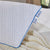 A close-up of the La-Z-Boy Serene Foam Density Pillow Traditional | Soft-Medium shows its white surface with zigzag patterns and blue piping. "LAGO" and "SOFT WASH" tags are visible as it rests on a cream knit blanket near a window with greenery.