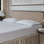 A neatly made bed with a beige headboard features two white textured pillows, one being the La-Z-Boy Serene Foam Density Pillow (Soft-Medium), and a matching white mattress. A lamp and book sit on the bedside table; beige curtains and wall art complete the scene.