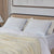 A neatly made bed features two La-Z-Boy Serene Foam Density Pillows (Soft-Medium) in beige cases, a cream and beige knit blanket, white sheets, a tan upholstered headboard, neutral curtains, a bedside lamp, and large artwork.