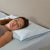 A woman with long brown hair sleeps peacefully on her side, resting her head on a La-Z-Boy Serene Foam Density Pillow Traditional | Soft-Medium, featuring a moisture-wicking cover in a calm bedroom with a textured white sheet and beige headboard.
