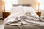 A cozy, unmade bed with white pillows and sheets is accented by the Herringbone Weave Blanket from American Blossom Linens. The softly lit room features wooden nightstands with lamps, a candle, and sunlight streaming in through two windows.