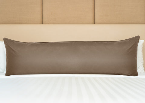 A long brown Pillowtex Body Pillow Cover rests on a neatly made bed with white linens against a cream-colored, padded headboard. The pillow is centered, extending across the width of the bed.