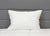 A JS Fiber "Ultra Down" Polyester Pillow | Firm stands upright against a gray headboard on a white, quilted comforter, adding soft texture and neutral tones. The JS Fiber pillow is machine washable for easy care.