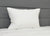 A JS Fiber "Ultra Down" Polyester Pillow | Firm sits upright on a white quilted mattress. A grey, padded headboard with horizontal seams creates a modern setting with crisp, machine washable bedding by JS Fiber.