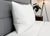 A neatly made bed features the JS Fiber "Ultra Down" Polyester Pillow | Firm, resting upright against a gray upholstered headboard with a white comforter. In the background, a wooden nightstand holds a potted plant beside a floor lamp with a white shade.