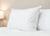 The Pillowtex Luxury Core Down and Feather Pillow (25% feather/75% down) by Pillowtex rests upright on a neatly made bed with matching white comforter, set against a beige wall and softly glowing bedside lamp for a serene atmosphere.