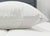A close-up of the Pillowtex Body Pillow Cover, made from antimicrobial copper-infused bamboo, shows its visible zipper on a white quilted surface. The minimal background includes part of a gray headboard and white wall for a clean, modern look.
