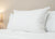 The Pillowtex Luxury Core Down and Feather Pillow (25% feather/75% down) by Pillowtex rests upright on a neatly made bed with white sheets, a beige wall, and a glowing lamp creating a cozy, inviting bedroom atmosphere.