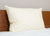 The Down Etc. Aquaplush Pillow, a large, cream-colored hypo-allergenic pillow with a smooth surface, rests upright against a brown wooden headboard on a white mattress, set against a light beige wall for a clean bedroom look.