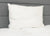 The Pillowtex High End White Goose Down Pillow | Soft Support rests upright on a white mattress against a gray upholstered headboard. Ideal for stomach sleepers, this minimal, well-lit scene shows visible tags on the pillow’s side.