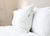 A neatly made bed features two white pillows stacked against a white headboard. The front pillow is the Pillowtex Luxury Core Down and Feather Pillow (25% Feather/75% Down) by Pillowtex, both resting on a smooth white comforter.