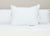 A close-up of a neatly made bed shows three large Hollander Eco-Smart Down Alternative Pillows made in the USA, arranged side by side against a light beige wall. The crisp white pillowcases highlight the eco-friendly, recycled polyester fill.