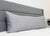 A large gray body pillow with a Pillowtex Body Pillow Cover | Antimicrobial Copper Infused Bamboo rests against a modern gray headboard on a neat white bed. A small green potted plant appears near the bed’s right edge.