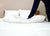 A hand presses into a Pillowtex Luxury Core Down and Feather Pillow (25% feather/75% down) on a neatly made bed with white bedding, wooden headboard, two plush pillows in back, and an alarm clock on the bedside table.
