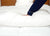 Wearing dark blue pajamas, a person presses their hand into the Creative Bedding Fossfill Pillow | Antibacterial, highlighting its soft, synthetic down feel. A wooden headboard and another pillow are visible in the background.