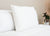 The Pillow Factory Housekeeper's Choice Heavenly Spiral Pillow—featured at many Borgata Hotel Casino & Spa®—stands upright on a pristine bed with a white comforter; in the background, a lush potted plant sits atop a table against a beige wall.