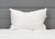 The Pillowtex High End White Goose Down Pillow, offering medium-firm support, is propped against a gray upholstered headboard with a white quilted comforter, giving the bed a soft, neat, and inviting minimalist look.