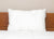 The Pillowtex Like Down Pillow by Pillowtex, a white hypoallergenic pillow with a fabric tag, sits upright against a wooden headboard on a neatly made bed with white bedding and a plain beige wall in the background.
