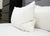 A close-up of a neatly made bed with white sheets and a Down Etc. Rhapsody Wrap Down/Feather Pillow—featured at many Hyatt® Hotels—propped against a gray headboard, with a black dresser and books on shelves softly blurred in the background.