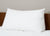 The Pillowtex Reading Wedge Bed Pillow by Pillowtex sits upright against a wooden headboard on a neatly made bed with a white comforter, while light beige walls provide a minimal and clean bedroom setting.