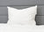 A Pillowtex High End White Goose Down Pillow with medium-firm support sits upright against a gray headboard on a bed with a white, quilted comforter, creating a minimal, fresh look for a comfortable bedroom.