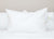 A Manchester Mills Down Dreams Classic Firm Pillow (formerly Classic Too) with crisp edges stands upright on a white quilted bedspread. A white and a yellow tag are attached to the pillow’s right side, in front of a plain light beige wall.