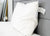 The Pillowtex High End White Goose Down Pillow | Soft Support rests upright against a light gray headboard on a tidy bed—perfect comfort for stomach sleepers. A bedside table and gold-accented clock add to the serene scene.