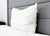 The Pillowtex High End White Goose Down Pillow with medium-firm support rests against a tall gray headboard on a neatly made bed with a white comforter, with part of a wooden nightstand visible in the background.