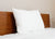 A single large, fluffy Pillowtex Like Down Pillow by Pillowtex rests against a wooden headboard on a neatly made bed with a white comforter, set against a plain beige wall. The minimal scene features soft lighting and a tidy atmosphere.