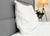 A Pillowtex High End White Goose Down Pillow with soft support by Pillowtex rests upright against a gray headboard on a neatly made bed, while a floor lamp and a potted plant create a calm, minimalistic scene.