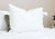 A Manchester Mills Down Dreams Classic Soft Pillow, featured at many hotels, sits upright on a white bedspread with a tag attached. Partially behind it is a smaller pillow with a green leafy pattern, and a clock appears on the neutral background.