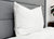 A Pillowtex High End White Goose Down Pillow with medium-firm support stands upright against a gray upholstered headboard, complemented by white bedding and a wooden nightstand with a potted plant for a clean, modern look.