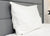 The Pillowtex High End White Goose Down Pillow | Soft Support rests upright against a gray headboard on a neatly made bed with a white comforter, with a dark-leafed plant in the background. Ideal comfort for stomach sleepers.