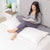 A woman with long dark hair, in a gray top and leggings, reads on a neatly made bed. She is comfortably supported by the Pillowtex White Goose Feather and Down Body Pillow by Pillowtex, featuring a stylish cover.