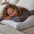 A woman with curly hair and a gray top lies on her stomach on a bed, looking comfortable as she rests her head and hands on a plush La-Z-Boy Fiber Density Pillow by La-Z-Boy in a softly lit bedroom with neutral bedding.