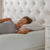A woman with curly hair lies on her side on a La-Z-Boy Fiber Density Pillow, finished with gold trim. She looks relaxed in a sleeveless gray top, resting on a bed with a textured mattress and tufted headboard.