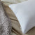 A La-Z-Boy Fiber Density Pillow with a "FIRM" tag rests on beige bedding beside a chunky gray knit blanket, while part of a light quilted blanket appears in the background for a cozy, inviting look.