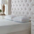 A neatly made bed with a white, textured mattress, La-Z-Boy Fiber Density Pillows, and a tall tufted beige headboard. A white nightstand with a lamp, potted plant, and decor sits nearby in this bright, serene setting.