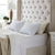 A neatly made bed with a tall, tufted beige headboard, white sheets, and three large La-Z-Boy Fiber Density Pillows. White nightstands flank the bed; one holds a lamp with a beige shade and a potted plant. Sunlight streams in.