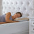 A woman with curly hair smiles as she sleeps on her side, resting her head on a La-Z-Boy Hybrid Bliss Memory Foam | Richloft Fiber Foam Core Pillow. The bed has a white tufted headboard, and a bedside table with drawers sits nearby.