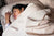 A woman with dark hair sleeps peacefully, her head resting on Colored Cotton Bed Pillowcases by American Blossom Linens—crafted from natural heirloom cotton and American made—creating a calm, comfortable scene in a softly lit bedroom.