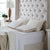 A neatly made bed with a tall, tufted beige headboard features two white pillows and two beige-trimmed wedge pillows, including the La-Z-Boy Hybrid Bliss Memory Foam Pillow. A bedside lamp and plant are placed near the sunlit window.